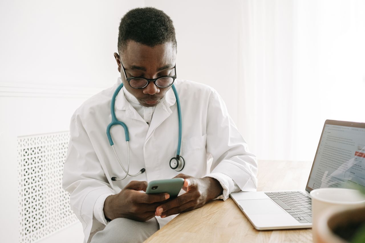 services-04 Doctor in a medical office checking a smartphone with a laptop on the table, wearing eyeglasses and stethoscope.
